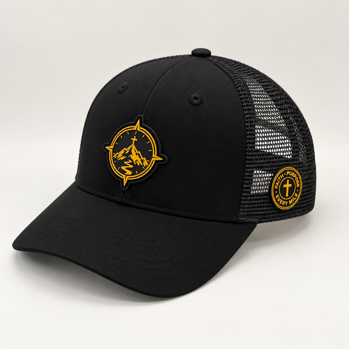 A black trucker-style baseball cap with a curved brim, black mesh back panels, and two circular embroidered patches in yellow and black. One patch features a mountain and compass design on the front, and the other is on the side mesh.
